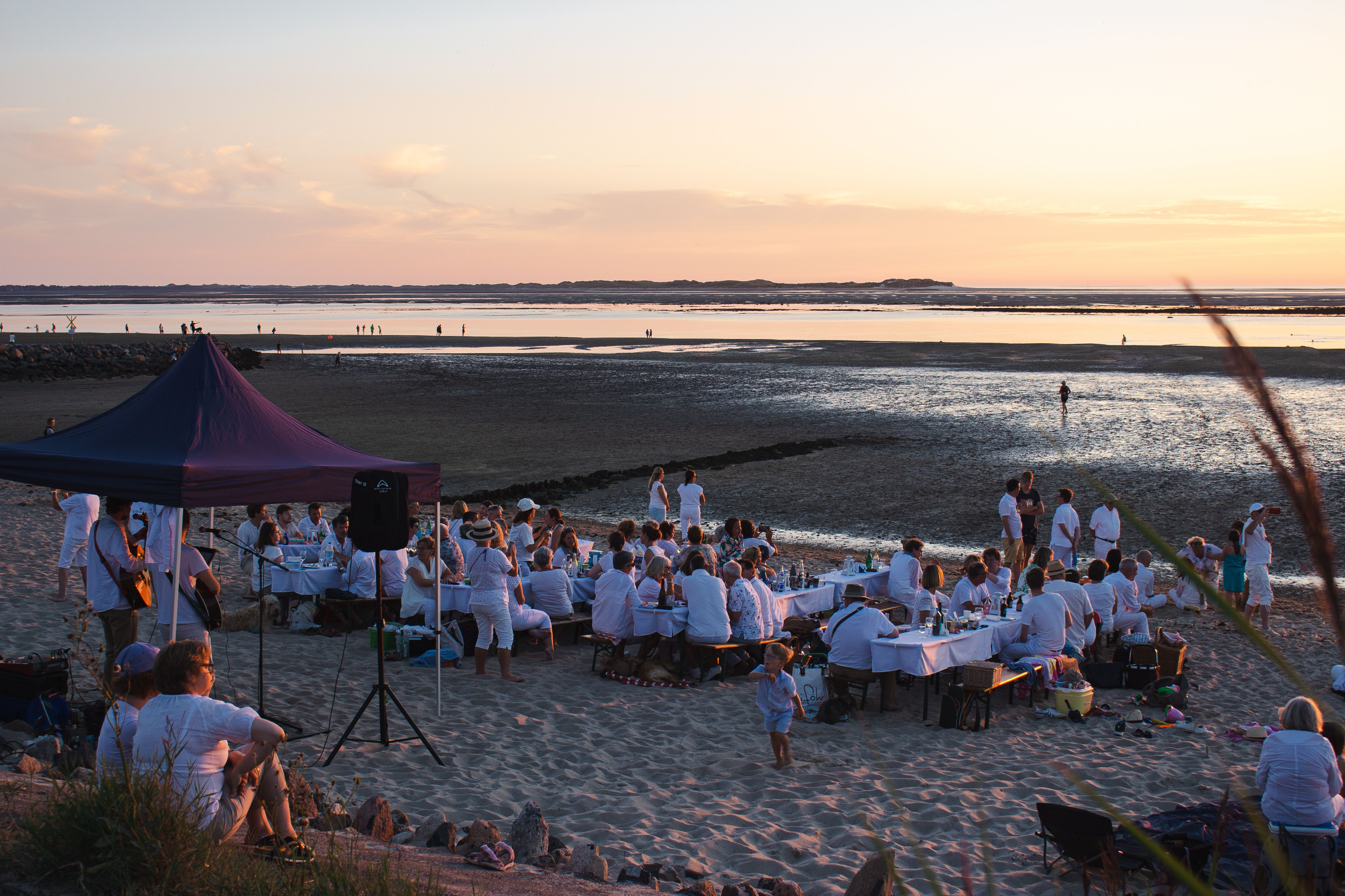 Teilnehmnde des White Dinners m Utersumer Strand auf der Nordseeinsel Föhr genießen den Sonnenuntergnag bei Live-Musik, Picknick und Gesellschaft