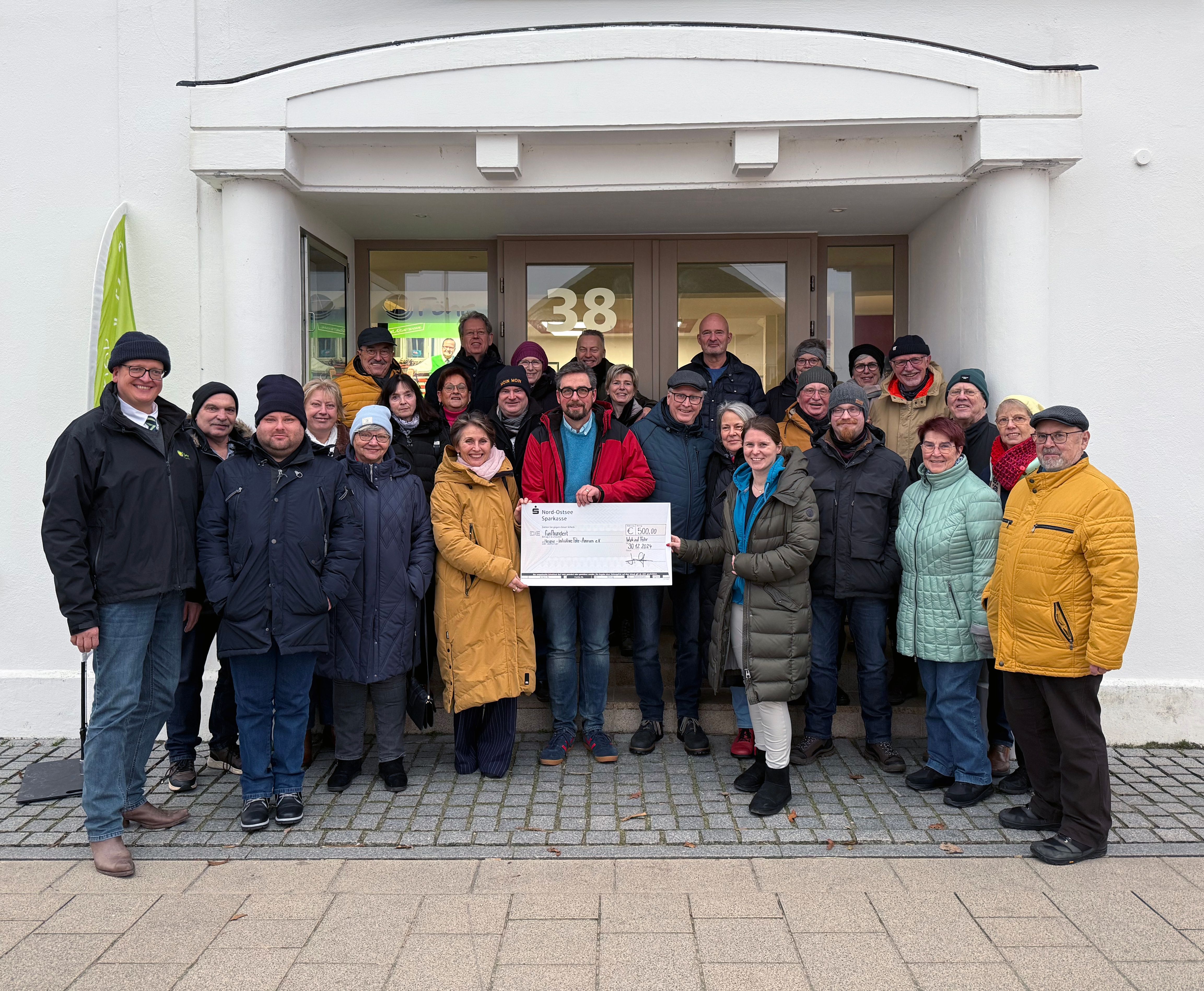 Gruppe von Menschen überreicht symbolischen Scheck vor einem weißen Gebäude.